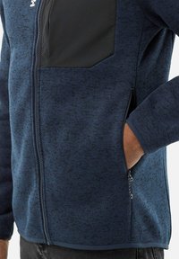 Veste en polaire bleu foncé avec un panneau noir sur la poitrine, des poches latérales, des poignets côtelés et un motif de tissu texturé.