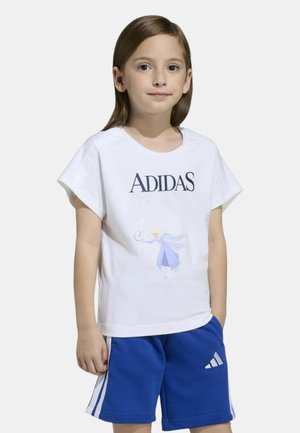 Bambino piccolo che indossa una maglietta bianca Adidas con una grafica di principessa e pantaloncini Adidas blu con strisce bianche, in piedi con le mani rilassate.