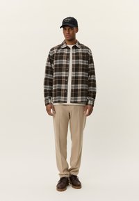 Braun-weiße karierte Flanellbluse, darunter ein cremefarbenes Hemd, beiges Tuch, braune Schuhe und eine schwarze Mütze mit weißem Text.