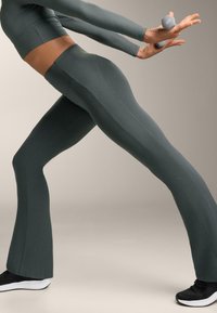 Tamnozeleni uski tajice s rebrastom teksturom, proširenim dnom i bočnim šavovima, uparene s crnim sportskim cipelama i sivom bučicom.