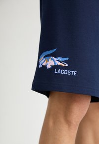 Shorts din bumbac de culoare bleumarin, cu un logo mare, stilizat de crocodil în nuanțe de albastru și roz, și textul "LACOSTE" în alb, pe marginea inferioră.