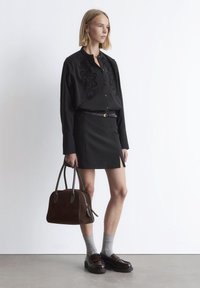 Woman wearing black embroidered blouse and matching skirt, holding a brown leather handbag, standing on gray floor against white wall.