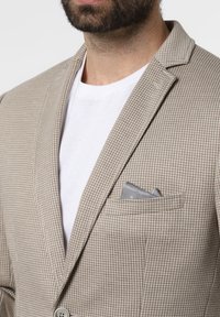 Beżowa marynarka w wzory z wyciętymi klapami, zapinana na jeden guzik, z szarą poszetką w kieszonce na piersi, noszona na białym t-shircie.