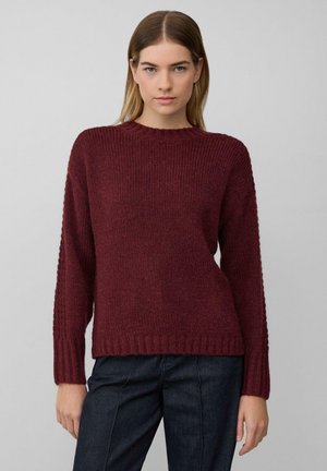Vrouw in een bordeauxrode gebreide trui met lange mouwen en een donkere spijkerbroek, staande tegen een effen lichtgrijze achtergrond.