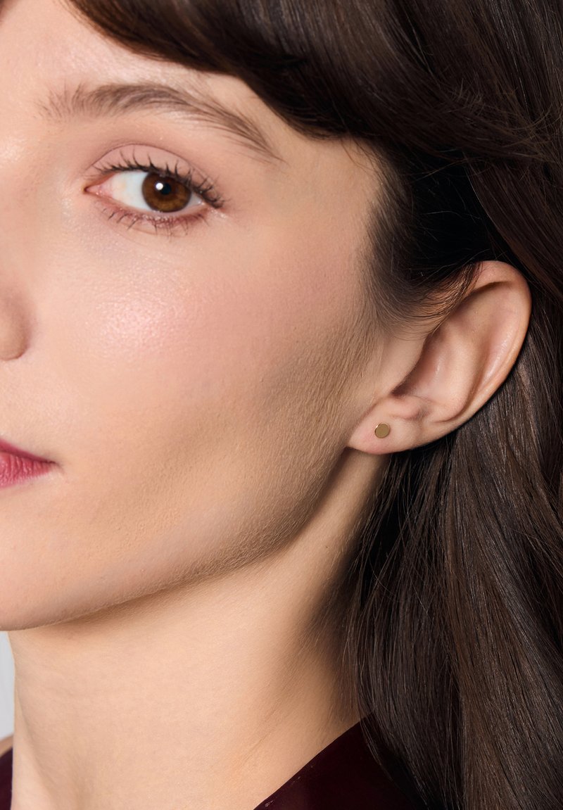 Złoty kolczyk w kształcie minimalistycznej, okrągłej formy, założony na uchu modelki z gładką skórą i naturalnym makijażem.