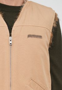 Beżowa kamizelka z miękkiego materiału, z zamkiem z przodu, kieszenią po bokach oraz brązowym futerkiem wzdłuż szwów na ramionach. Widoczna ciemnozielona warstwa pod spodem.