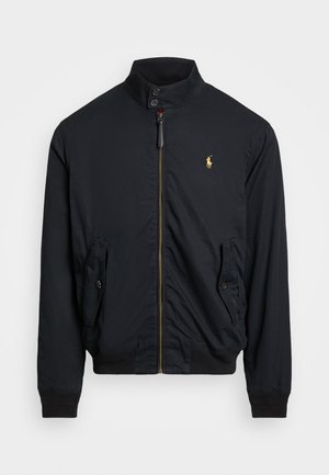 Svart nylonjakke med høy krage, gullglidelås, to sidelommer med knappklaffer og en liten brodert logo på brystet.