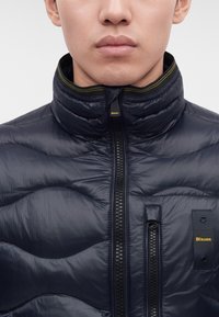 Homme portant une veste matelassée bleu marine avec col haut et logo Blauer visible sur la fermeture éclair et la poche poitrine.