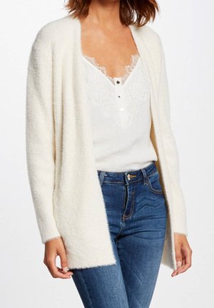 Femme portant un cardigan blanc pelucheux par-dessus un caraco blanc bordé de dentelle, associé à un jean bleu, debout devant un fond uni.