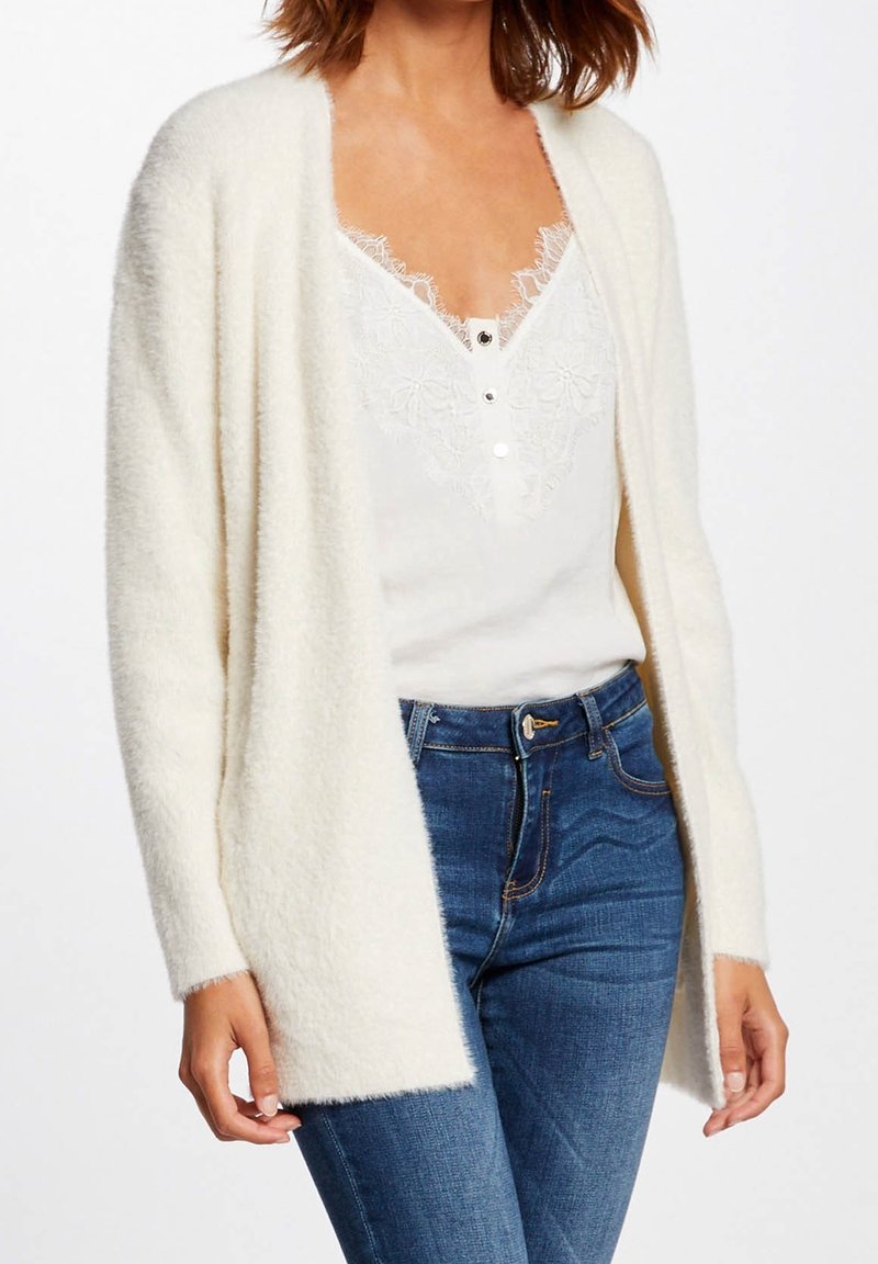 Femme portant un cardigan blanc pelucheux par-dessus un caraco blanc bordé de dentelle, associé à un jean bleu, debout devant un fond uni.