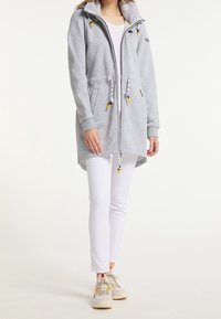 Žena oblečená v světle šedé mikině s kapucí a bílými šňůrkami, bílé těsné kalhoty a vícebarevné tenisky, stojící na bílém pozadí.