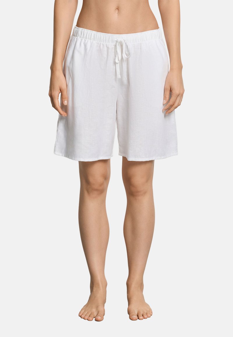 Femme portant un short blanc ample à la taille ajustable, arrivant au genou, debout pieds nus devant un fond uni.