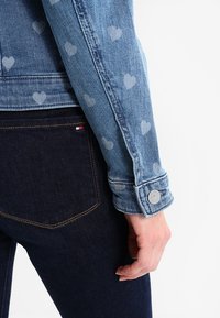 Ljusblå denimjacka med hjärt-mönster, utrustad med knappmanschetter och synlig sömnad, i kombination med mörkblå jeans.