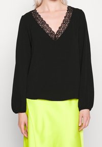 Blouse noire à manches longues avec col en V en dentelle, associée à une jupe en satin jaune néon vif portée par un mannequin blond.