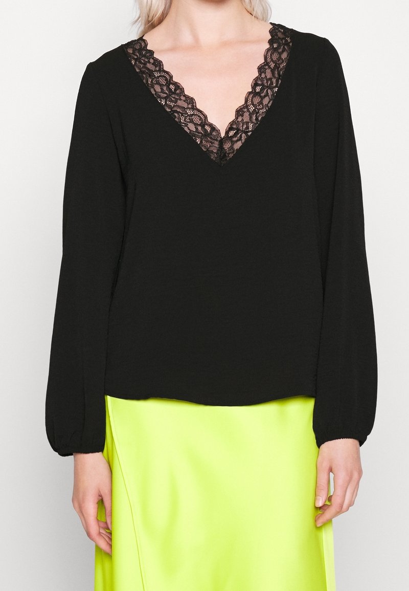 Blouse noire à manches longues avec col en V en dentelle, associée à une jupe en satin jaune néon vif portée par un mannequin blond.