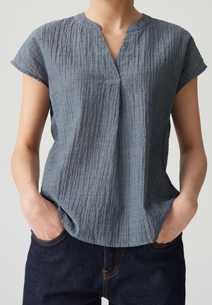 Personne portant un chemisier bleu-gris texturé à manches courtes avec un col en V, mains dans les poches d'un jean en denim bleu foncé.
