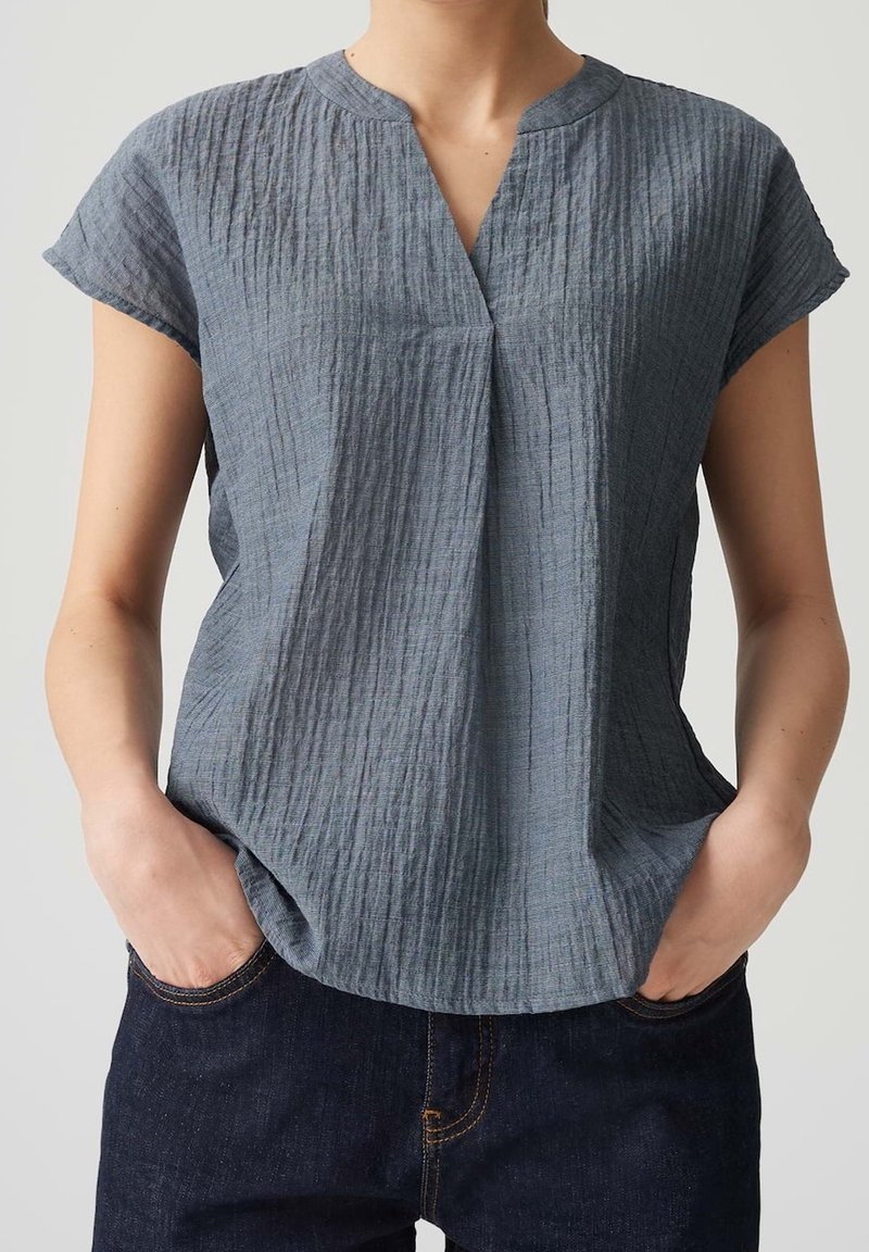 Personne portant un chemisier bleu-gris texturé à manches courtes avec un col en V, mains dans les poches d'un jean en denim bleu foncé.