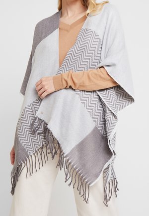 Femme portant un haut beige à manches longues et un pantalon de couleur claire, drapée d'un poncho gris et blanc à franges avec un motif en zigzag.