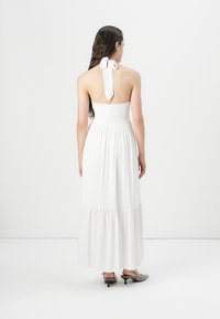 Hvit, ermeløs maxikjole med strikket liv og lagdelt skjørt. Har en halter neck-bånd. Modellen har på seg grå hælesko.