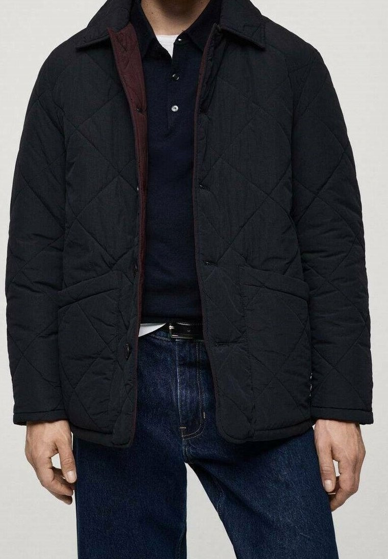 Homme portant une veste noire matelassée sur une chemise bleu marine, associée à un jean bleu foncé et une ceinture noire, debout les mains détendues.