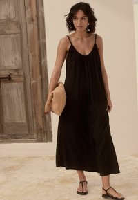 Robe noire au design fluide, avec des fines bretelles et un tissu texturé. Accordée avec une pochette beige et des sandales noires. Fond neutre.