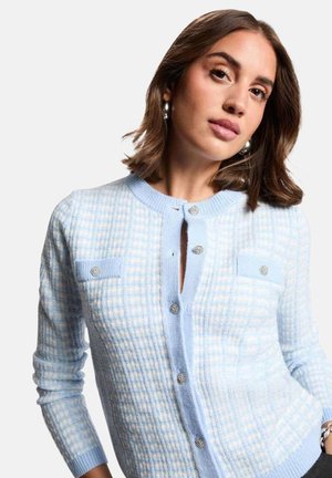 Femme aux cheveux bruns mi-longs portant un cardigan à carreaux bleu clair et blanc avec des boutons scintillants et des créoles argentées.