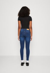 Jeans skinny de ganga azul com uma cintura alta, bolsos traseiros e um rótulo de pele castanha na cinta. Modelo a usar uma blusa preta e sapatos.