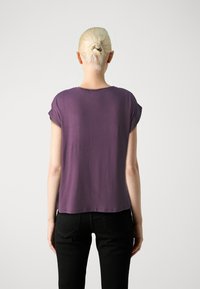 Top viola a maniche corte con vestibilità rilassata, texture liscia e scollatura rotonda, abbinato a jeans neri visibili in vita.