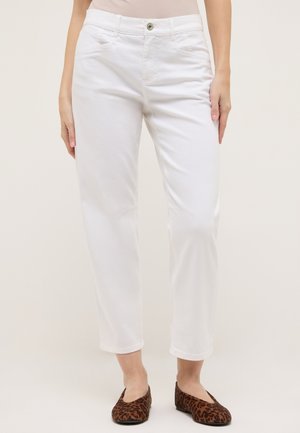 Frau trägt weiße, knöchellange Hose und braune, flache Schuhe mit Leopardenmuster, steht vor einem schlichten, hellen Hintergrund.