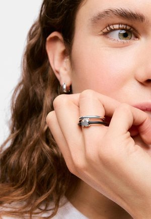 Primer plano de una persona con cabello rizado que lleva un anillo de plata con un colgante en forma de estrella y pendientes de aro plateados a juego.