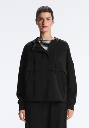 Femeie purtând un hanorac negru peste un tricou în dungi alb-negru, stând pe un fundal deschis, simplu.