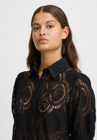 Camicia nera con bottoni caratterizzata da intricati motivi in pizzo, con colletto strutturato e maniche lunghe. Materiale leggero con una finitura testurizzata.