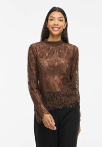 Femme souriante portant un chemisier à manches longues en dentelle florale marron et un pantalon noir, debout devant un fond blanc.