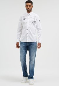 White long-sleeve shirt with black buttons and printed text, paired with light blue jeans and white sneakers, worn by a male model.