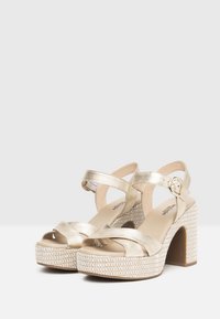 Sandales à plateforme métalliques dorées avec des brides croisées et une boucle décorative. Semelle intermédiaire tissée texturée et talon épais. Doublure intérieure lisse.