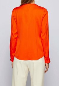 Långärmad blus i klar orange satin med rak klippning, rundad nederkant och knäppte manschetter, bärs över krämfärgade byxor.