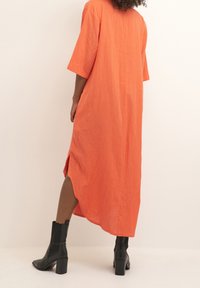 Pitkä, oranssi mekko lyhyillä hihoilla, rento malli, sivuhalkiot ja kevyttä materiaalia, yhdistetty mustiin korollisiin saappaisiin.