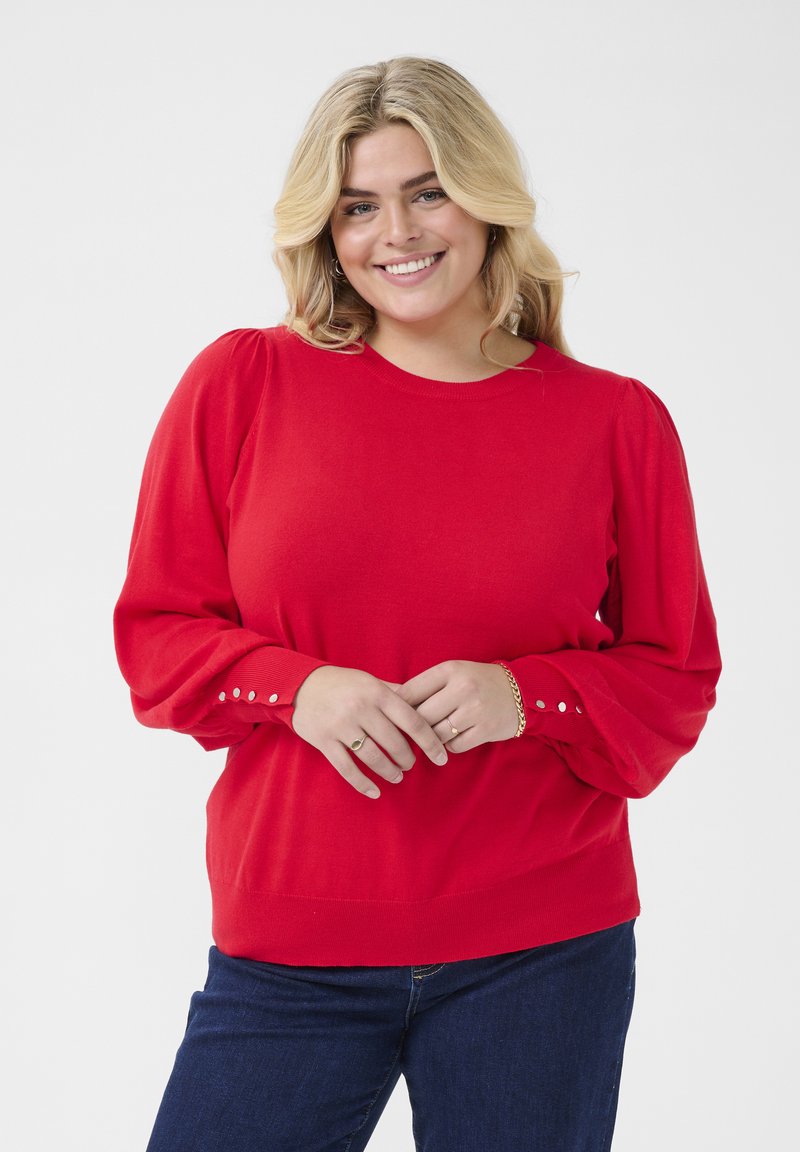 Pull en tricot rouge avec de longues manches bouffantes ornées de boutons à l'avant-bras, un col rond et une coupe décontractée. Porté par-dessus un jean en denim foncé.