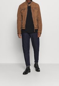 Uomo in giacca da motociclista in suede marrone, maglietta nera, jeans blu scuro e stivali alla caviglia di pelle neri in piedi su un pavimento grigio.