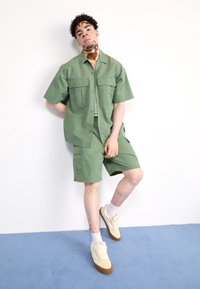 Grön kortärmad skjorta och matchande shorts i lätt tyg, med två bröstfickor. Beigea sneakers med ljusa strumpor kompletterar outfiten.