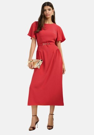 Femme en robe midi rouge à manches courtes avec ceinture, tenant une pochette dorée, portant des boucles d'oreilles dorées, un bracelet et des sandales noires à talons hauts.