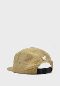 Vaaleanruskea lippalakki, jossa on kaareva lippa, säädettävä musta hihna ja pieni valkoinen brodeerattu logo takana. Teksturoitu kangas, rento muotoilu.