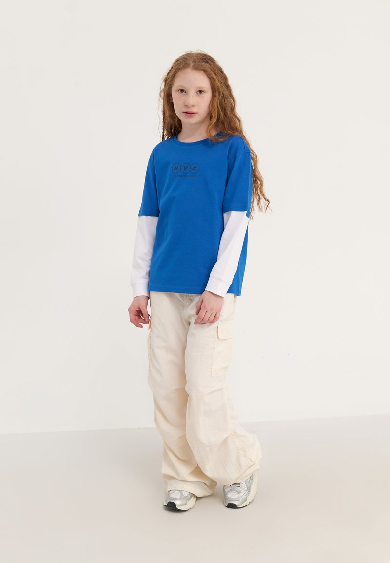 Blue t-shirt with white long sleeves, featuring black text. Paired with cream cargo pants and silver sneakers.