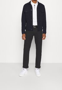 Chaqueta vaquera azul marino sobre un polo blanco, combinada con pantalones oscuros y zapatillas blancas. Cuenta con dos bolsillos en el pecho y cierre de botones.