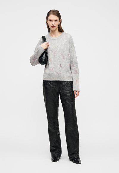 Γυναίκα με ανοιχτόγκρι πουλόβερ με διακριτικά σχέδια, μαύρο παντελόνι με φαρδιά πόδια και υφή, μαύρα μυτερά παπούτσια, κρατώντας μια μαύρη τσάντα ώμου.