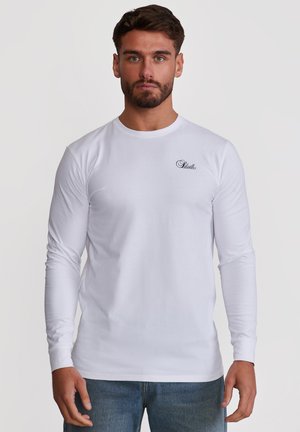 Uomo con barba che indossa una maglietta bianca a maniche lunghe con un piccolo logo nero a scritta sul petto, abbinata a jeans blu, in piedi davanti a uno sfondo bianco.