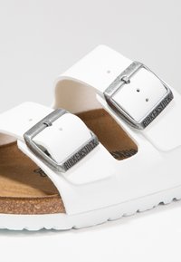 Sandali Birkenstock bianchi con due fibbie metalliche regolabili, soletta in sughero e suola in gomma texturizzata. Design classico con punta aperta.