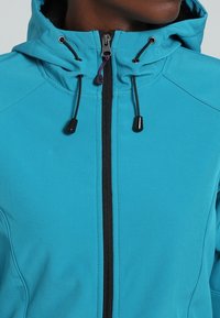 Personne portant une veste à capuche bleu sarcelle avec une fermeture éclair noire et des cordons de serrage noirs réglables autour de la capuche.