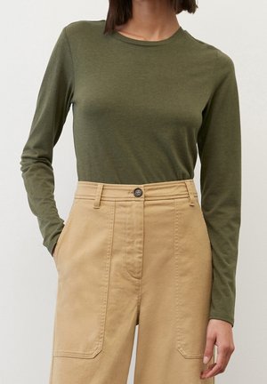 Frau trägt ein olivgrünes, langärmliges, eng anliegendes Oberteil, das in hoch taillierte beige Hosen mit aufgesetzten Taschen vorne und Knopfverschluss gesteckt ist.