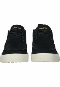 Zapatos de ante negros con puntera redondeada, que presentan cordones negros y una suela de goma blanca. La marca en dorado en la lengüeta añade un detalle distintivo.